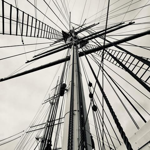 A black and white photograph looking up at the mast of a tall ship, featuring rigging, ropes, and a lattice ladder against a cloudy sky.