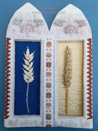 My Shrine Of Ceres, part of the Biodiversity Shrine Project. A two-part shrine in the shape of Gothic church windows, built three-dimensionally from cardboard painted white with gesso. On the left, an embroidered ear of grain on blue fabric. On the right, a real one on golden fabric. Decorated with folklore braids and showing traditional Ukrainian embroidery patterns with tree-of-life symbols.