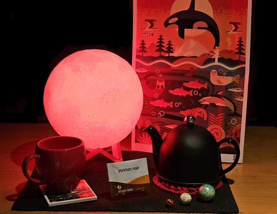 Tea tableau commemorating March 2025 total eclipse. There is a luminous red moon, three marbles signifying Sun, Earth, and Moon, and a backdrop that is a dramatically lit poster of an orca breaching before a full moon with dozens of glowing sea creatures below. A fat round red mug sits upon a coaster tile that matches the poster. A black teapot is beside it on a red and black crocheted mat.The tea is Yunnan Noir black tea from Adagio.