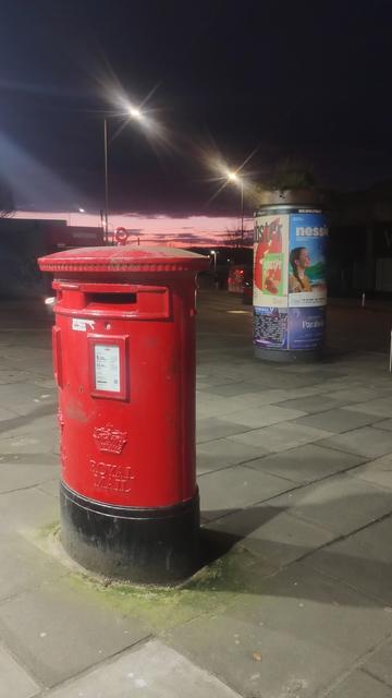 Red British Royal Mail post box at night, lit by bright light from a nearby convenience store, with a splash of red on the horizon of the twilight sky behind it
