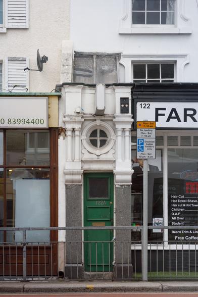 Sandwiched between two shops in a row of terraced buildings, an incredibly narrow entrance with grey granite pillars, white-painted columns and other ornate architectural details including a circular window, and a small, narrow, green-painted door with brass fittings and 122A numerals screwed on.