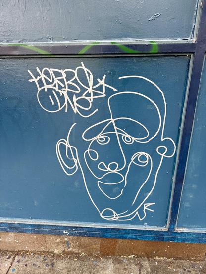 Silver paint marker graffiti on a green wall. It is a portrait drawing made in one long and squigly line. The artist's name appears to be "Herbs One", but I'm not sure on that.