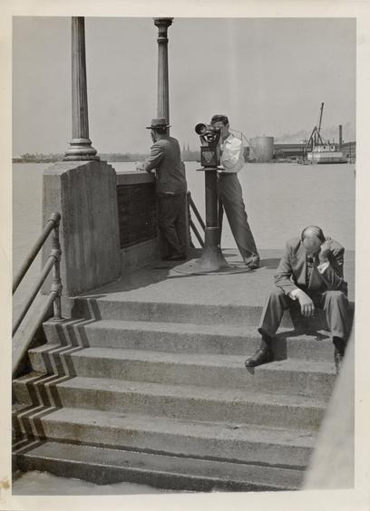 Black and white photo of three men on a set of stairs. One is looking out over the flooded city. Another peers through binoculars. A third is sitting on the steps and looking a little miserable. This is a flood in 1948.