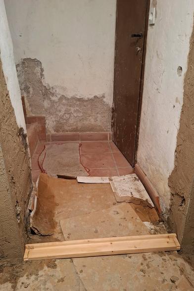 The image shows ongoing renovation work in a narrow passage with an aged brown door at the end.  The floor is partially covered with protective cardboard sheets, suggesting active construction. The rough, unfinished walls and visible repairs indicate a working place. A wooden piece is placed on the cardbords as a the lower individual strip for the door frame.