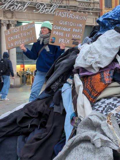 Devant le tas de vêtements, une activiste montre les pancartes "la fast-fashion entretient la précarité" et "quelqu'un, quelque part, paye le vrai prix"