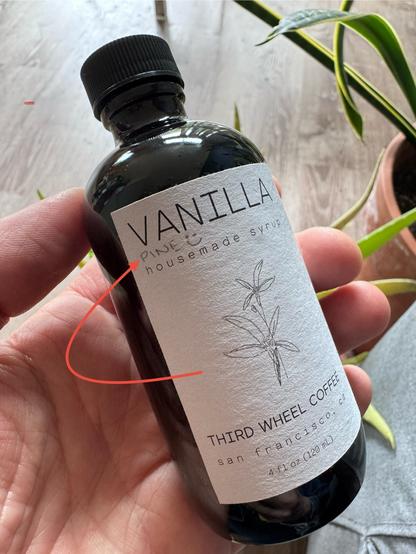 A bottle of syrup with a label that says:

VANILLA
PINE
housemade syrup
THIRD WHEEL COFFEE san francisco.
4 f1 0z (120 mL).

The word “pine” has been added with pencil.