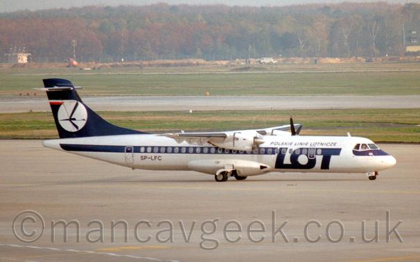 Side view of a white, high-winged, twin propellor-engined airliner taxiing from left to right.
There is a thick blue stripe running along the body, covering the passenger cabin windows, with large blue "LOT" titles on the forward fuselage, and smaller "Polskie Linie Lotnicze" above that logo.
The tail is the same dark blue, with a white circle containing a stylised blue flying bird.
Concrete apron fills the foreground, with large areas of grass either side of a grey runway leading up to trees in the distance, under hazy grey sky.