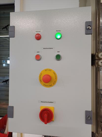 An electrical control cabinet with a gray metal door. On the door, there are several controls: 

at the top, a red fault indicator light and a green operating light (which is illuminated). Below them, two buttons for exhaust ventilation—one red for "OFF" and one green for "ON." In the middle, there is a large emergency stop button with a red knob and a yellow background. At the bottom, a main switch with a red rotary handle and a yellow safety cover.

The control cabinet is mounted on a wall.
