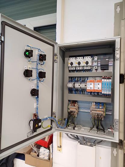 The image shows the inside of a wired control cabinet.

On the inner side of the door are the contacts for push buttons and signal lights, neatly wired and secured. Inside the cabinet, terminal blocks, motor protection switches, airflow monitors, zener barriers, and a 24V DC power supply are organized and connected on DIN rails with cleanly routed wiring.