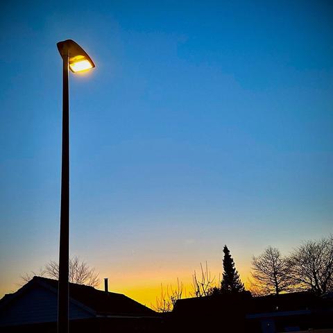 A dimly lit streetlamp stands against a clear blue sky at dusk, with a colorful sunset featuring shades of orange and yellow. Silhouettes of trees and rooftops are visible in the foreground.