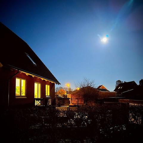 A serene nighttime scene featuring a house with lit windows, under a clear blue sky illuminated by a bright moon. Nearby structures and trees are visible, along with soft street lighting.