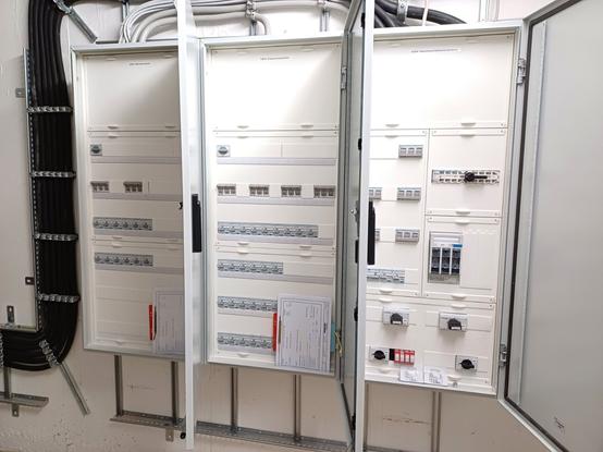 Three open electrical distribution cabinets in a technical room, with white plastic covers and multiple rows of circuit breakers, residual current devices, and other electrical components. The right cabinet is labeled 'UPS main distribution' and contains switches, disconnectors, and other electrical components. On the left, thick black cable bundles are secured to cable trays.