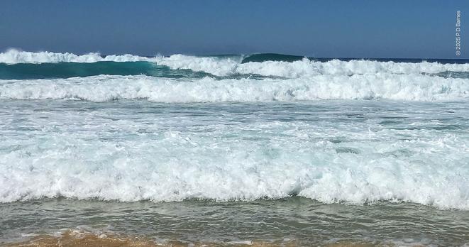 Low view of lines of small waves breaking and coming in to shore.  The image is predominantly the bright white of sea-foam from the waves, which are running almost to the camera, with glimpses of clear green water beneath and within the swell. The sky is a clear, dull blue.