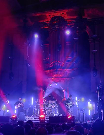 A band on stage, guitarists to the left and right and drummer in the middle. Red and blue lights bathe the stage and highlight the old.pipe organ looming behind them.