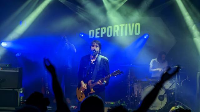 Chanteur-guitariste sur scène de concert.
Derrière il est écrit Deportivo