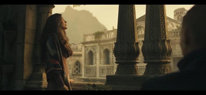 The image depicts a scene with Moiraine standing on a balcony, gazing outwards. She has long, wavy hair and is wearing a multicolored jacket over a light-colored dress. The balcony is adorned with ornate stone pillars featuring intricate carvings. In the background, there is a large, detailed building with arches and decorative elements, set against a mountainous landscape.