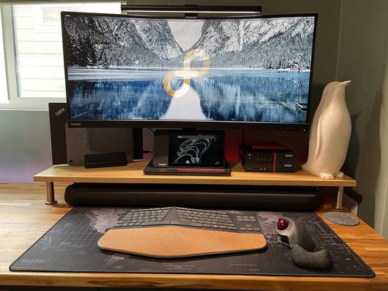 Desktop setup with a 34" curved ThinkVision monitor in the center on a monitor stand. It is flanked by a ThinkPad T480 and Lenovo Duet tablet to the left, a pair of ThinkCentre M900 Tinys and a penguin statue to the right. A BenQ light bar sits atop the monitor while a ThinkSmart Android display sits below. A Yamaha soundbar is under the monitor stand and a Lenovo keyboard and a Logitech Trackman Ergo sits in front on a classic IBM black and white world map deskpad. All of this rests upon a teak butcher block desk top.