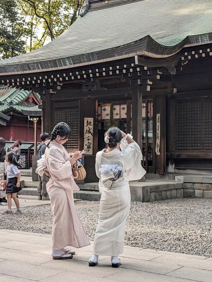 Two women in white kimono standing in front of a temple hall. The one on the right is taking a picture of the building, while the one on the left looks at her phone. There is a child on the left of the photo. The temple hall is made of wood and the floor is paved with clean cut stone.