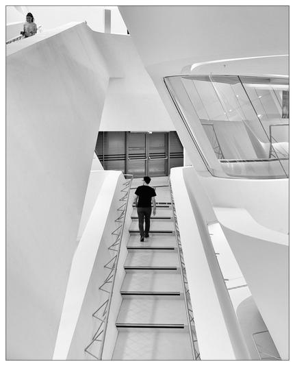 This image captures a modern architectural interior of a library. The photograph, taken in black and white, emphasizes the geometric shapes and the stark contrasts between the angular and curved elements of the structure. There are two persons featured: one descending a staircase and another observing from an upper level. The style and design suggest a focus on contemporary architecture, possibly emphasizing minimalism and the interplay of light and shadows.
