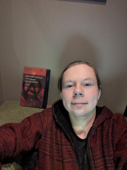 The individual is taking a self-portrait.

In the background, one can discern a book titled 'Rudolf Rocker: Anarcho-Syndicalism', which hints at their political views.

The foreground features a couch with a simple design, adding to the overall impression of a basic, unadorned environment.