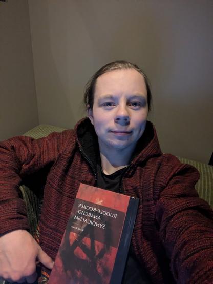 The individual is taking a self-portrait.

In the individuals hand, one can discern a book titled 'Rudolf Rocker: Anarcho-Syndicalism', which hints at their political views.

The foreground features a couch with a simple design, adding to the overall impression of a basic, unadorned environment.