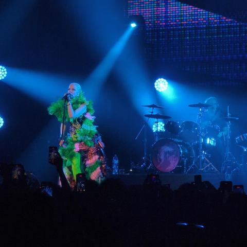 Shirley Manson vistiendo un llamativo atuendo de plumas verdes y rosas canta al micrófono en un escenario iluminado en azul. Detrás de ella, un baterista está sentado en una batería, parcialmente visible en la penumbra. La iluminación del concierto crea una atmósfera dramática. La multitud está presente, aunque solo se ven siluetas de personas sosteniendo teléfonos móviles.