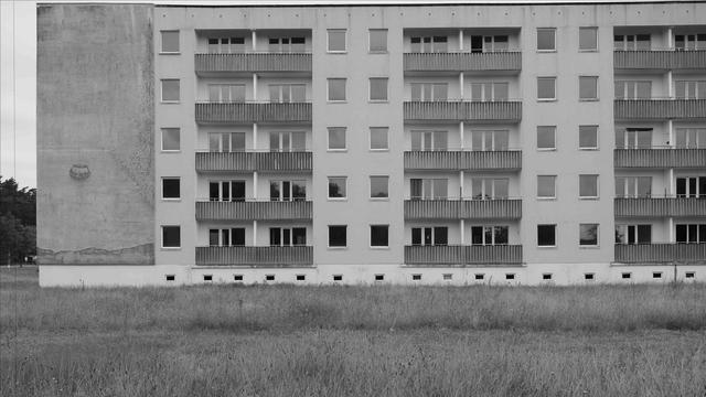 The image shows a black-and-white photograph of a large apartment building with multiple balconies. Most of the windows and balconies appear uniform and repetitive. There is an open grassy area in front of the building. The left side of the building has a bare, unpainted wall, which is in contrast with the rest of the structure.