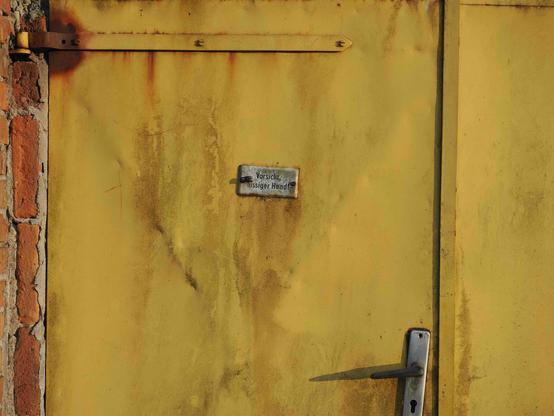The image shows a yellow metal door with rust stains. There's a small metal sign in the center. The door has a handle on the right and is set against a brick wall on the left. The sign reads "Vorsicht, bissiger Hund!" in German, which translates to "Beware of the dog!"