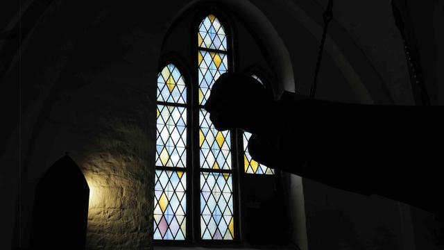 The image shows a silhouette of an object or figure against a backdrop of a stained glass window. The window has a traditional design with diamond-shaped panes in various colors. The scene is dimly lit, emphasizing the contrast between the dark silhouette and the illuminated window.