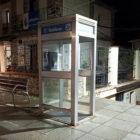 Telodescribot 

Una cabina telefónica de metal gris, parcialmente iluminada por la noche, se encuentra en una acera de piedra. En el interior, se ve un teléfono público azul. La cabina está ligeramente desgastada. Detrás de la cabina, se aprecia un edificio de piedra con un balcón y una valla metálica. Un banco de madera está situado en primer plano a la izquierda.