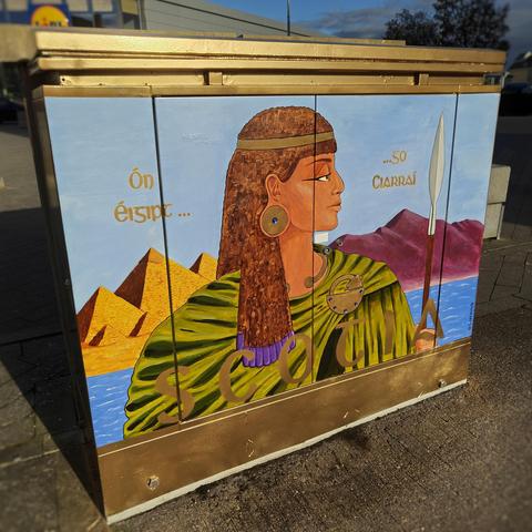 Photograph of a utility box depicting an Egyptian woman in Celtic ornaments and a green cloak, holding a spear. Behind her are the pyramids and the Sliabh Mis mountains, and the sea. It reads "Ón Éigipt...Go Ciarraí" and the name "SCOTIA"