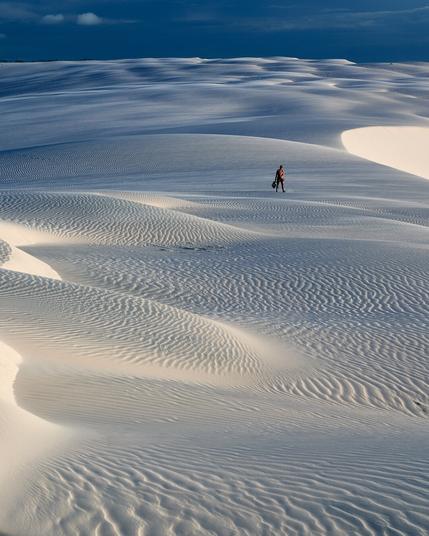 The photo shows a landscape of sand dunes until the horizon and a blue sky above. In the far there is a woman seen wandering alone through the dunes.