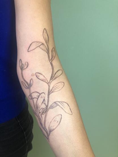 Tatouage d’une branche de sauge avec un petit personnage sur le bras
