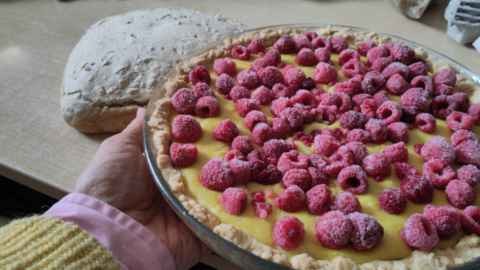 Une tarte aux framboises présentée devant une table beige avec un pain à la croûte beige clair. Les fruits roses parsèment une crème pâtissière jaune. Le plat à tarte est porté d'une main, pas si gauche que ça, sortant d'une manche de chemise rose dépassant d'un pull jaune à large maille.