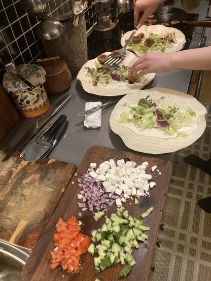 A kitchen counter with a cutting board with mixed vegetables on it, next to it, the hands of a person preparing falafel on bread.