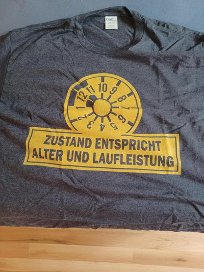 graumeliertes T-Shirt mit gelber TÜV-Plakette und der Schrift "Zustand entspricht Alter und Laufleistung.