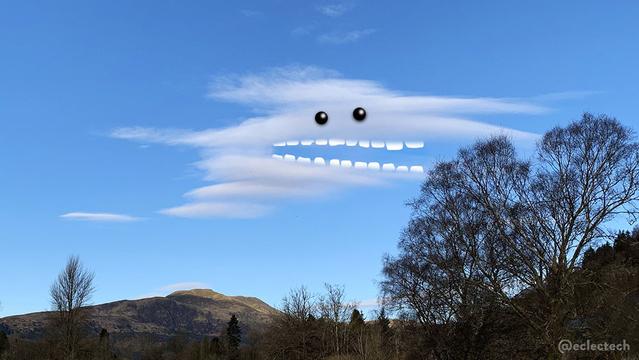 A photo of a mountain top, trees, and a big blue sky with one rather smooth but wibbly white cloud in the centre. The cloud looks like a number of fairly smooth clouds stacked together, and there is a V shaped gap in the middle which I have predictably added teeth to, then eyes above. It's looking very excited about the big, leafless tree on the right hand side.