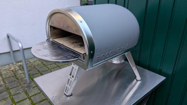 Ein Roccbox Gas-Pizza-Ofen von Gozney in der Farbe grau steht auf einem Tisch im Freien. Der Ofen hat eine Verlängerung zum Ablegen von Speisen und oder zum einfacheren Wenden der Pizzen. Im Hintergrund sieht man einen grünen Gartenschuppen. Der Ofen steht auf einem silbernen Metalltisch.