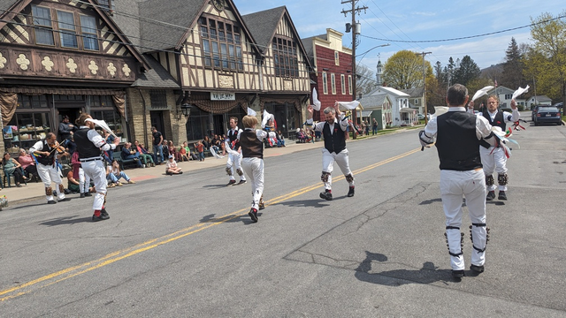 Six morris dancers (Bouwerie Boys Morris Dancers) are dancing for an audience in the street in front of an old building. The dancers and musician wear white shirts and trousers, black vests, red socks, black shoes, colored ribbons on black arm bands, and bells on the shins; they are holding white hankies which are raised overhead. Each has a red rose pinned to the left breast of the vest. The musician plays a fiddle. Photo taken in Gilbertsville, New York, April 2024.