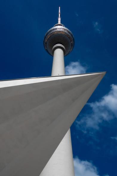 Der Fernsehturm in Berlin ragt imposant in den strahlend blauen Himmel, während das futuristische Dach im Vordergrund eine kantige Silhouette zeichnet. Die klare Architektur und die auffällige Perspektive verleihen dem Bild einen modernen, urbanen Charakter.