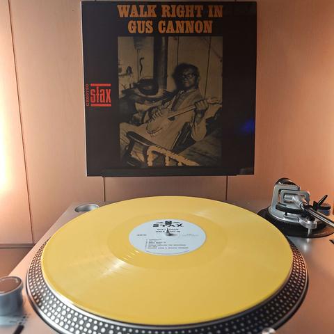 A yellow vinyl record sits on a turntable. Behind the turntable, a vinyl album outer sleeve is displayed. The front cover shows Gus Cannon sitting in a rocking chair holding a banjo.