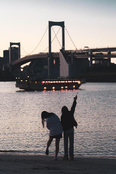 The image depicts two people near a body of water, engaging with bubbles, at either dusk. In the background, a boat adorned with lights is visible, with a large bridge and a cityscape in silhouette. The scene has a tranquil and picturesque ambiance.