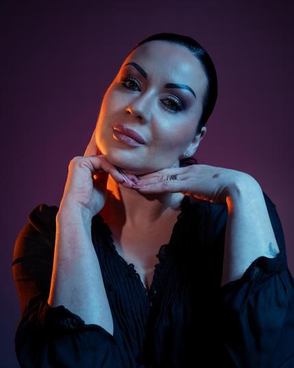 This image features a person posing with their hands under their chin, against a dark background. The lighting is artistic, with warm tones highlighting their face. The person is wearing a dark top and has neatly styled hair.