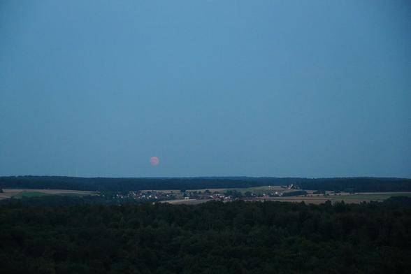 rotoranger Mond geht am blauen Himmel über einem kleinen Dorf auf, das zwischen Feldern und im Vorder- und Hintergrund Wäldern liegt.