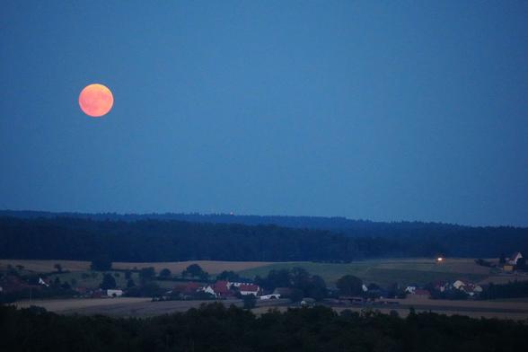 rotoranger Mond am blauen Himmel über einem kleinen Dorf zwischen Feldern und im Vorder- und Hintergrund Wäldern