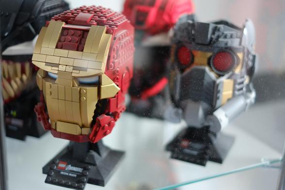A close-up image showcasing LEGO models of superhero helmets. The foreground features a LEGO Iron Man helmet, primarily red and gold in colour, displayed on a stand with a label that reads \"LEGO Avengers Iron Man.\" Next to it is another detailed LEGO helmet characterised by grey and gold colours with prominent red eyes, suggesting it is a different superhero character. The background shows parts of additional LEGO models. The items are displayed in a glass case.
