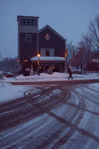 Vaguely Church-shaped building near a snowy crossroads in dusk light, tire tracks draw curves in the snow in the lower half of the picture.