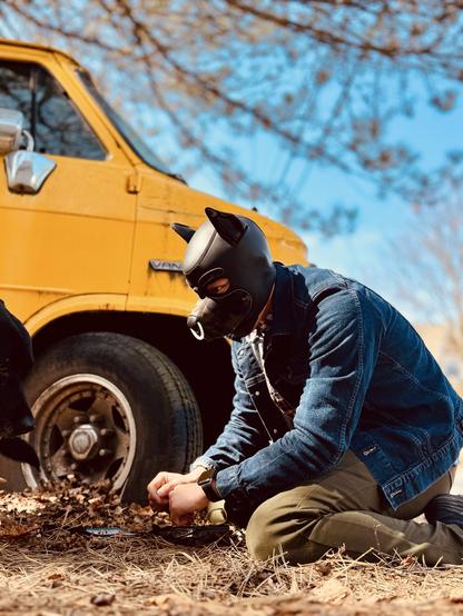 Sunlight streams through bare branches against a brilliant azure sky, casting a warm glow on this intimate moment captured in perfect detail. The  pup kneels attentively beside a vibrant mustard-yellow delivery truck, their paws extended to a black lab approaching from the left. The contrast between the vehicle's bold color and the figure's outfit is striking - a fitted denim jacket hugs shoulders while khaki pants meet the dried straw-covered earth. Most captivating is the glossy black/camo neoprene puppy hood adorning their head, complete with pointed ears and a white nose ring at the muzzle that catches the light brilliantly. The hood's smooth texture creates a sensual silhouette against the rustic backdrop, embodying devoted service and attentiveness as they work diligently on their task. The fallen pine needles and autumn detritus create a natural carpet beneath them.