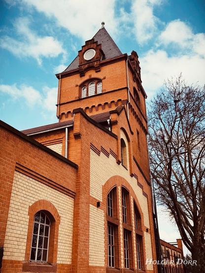 Das Bild zeigt den „Alten Schlachthof“ in Gießen, ein historisches Backsteingebäude mit einem markanten Turm. Der Bau besteht aus rötlichen und hellen Ziegelsteinen, die ein dekoratives Muster bilden. Der Turm hat ein steiles Dach mit einem kleinen Rundfenster und verzierten Fensterbögen. Der Himmel ist blau mit weißen Wolken, und rechts im Bild steht ein kahler Baum, der die Szenerie ergänzt. Die Architektur vermittelt einen industriellen, aber zugleich charmanten historischen Charakter.