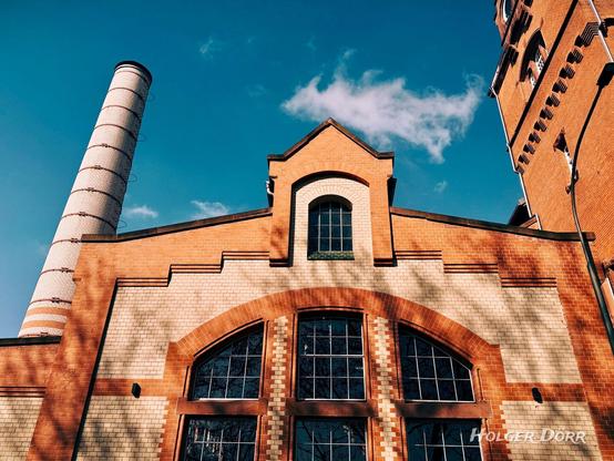 Die Fassade des „Alten Schlachthofs“ in Gießen, ein historisches Backsteingebäude mit großen Fenstern und kunstvollen Ziegelmustern. Im Hintergrund ragt ein hoher, weißer Industrieschornstein mit schwarzen Ringen in den blauen Himmel, während die Nachmittagssonne warme Schatten auf die Wand wirft. Rechts im Bild ist ein Teil eines Turms mit kleinen Fenstern und verzierten Backsteindetails zu sehen.